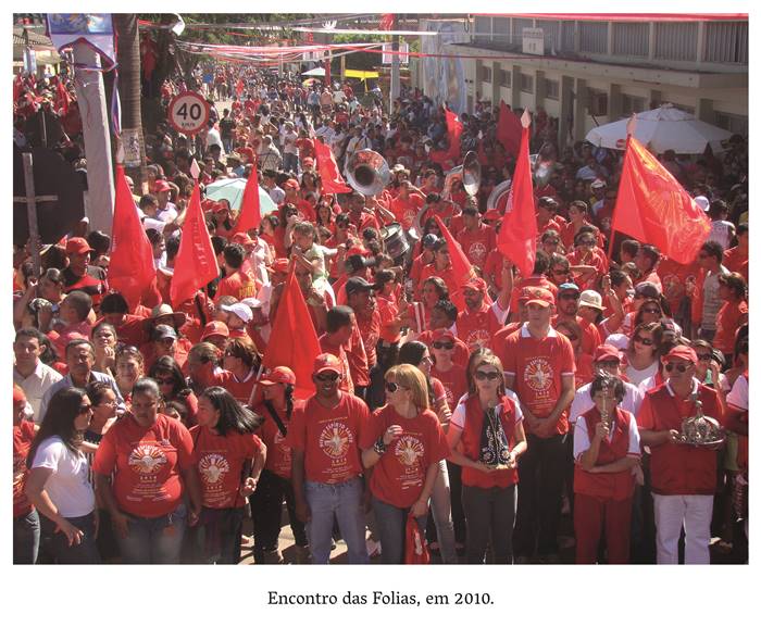  4 s- Encontro das Folias em 2010. Em espaço aberto e amplo homens, mulheres e crianças trajando camisetas vermelhas onde está escrito Divino Espírito Santo em dourado e branco. Erguendo bandeiras vermelhas caminham lado a lado em procissão. Em destaque, uma mulher branca de cabelos claros segura a imagem de Nossa Senhora de Aparecida (a imagem é negra coberta com manto azul escuro e dourado. Realização: Academia Planaltinense de Letras, Artes e Ciências (APLAC), pelo Ecomuseu Pedra Fundamental e pelo Coletivo Nativo Audiodescrição produzida  pelo Instituto de Promoção das Pessoas com Deficiência Visual Audiodescritora: Elma Lúcia Rodrigues Consultor: Fernando Rodrigues Este projeto é promovido com recursos do Fundo de Apoio a Cultura do DF